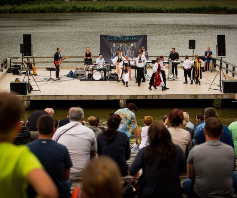 Traditional Romanian folk dancers and live band performing on an open air stage at the Astra Open Air Museum in Sibiu, Romania, with an audience watching from the lakeside
