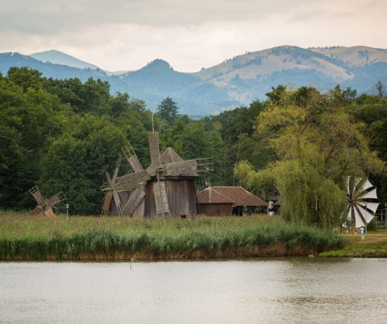 Traditional Romanian wooden windmills beside a lake at the Astra Open Air Museum in Sibiu, Romania, with the Carpathian Mountains in the background