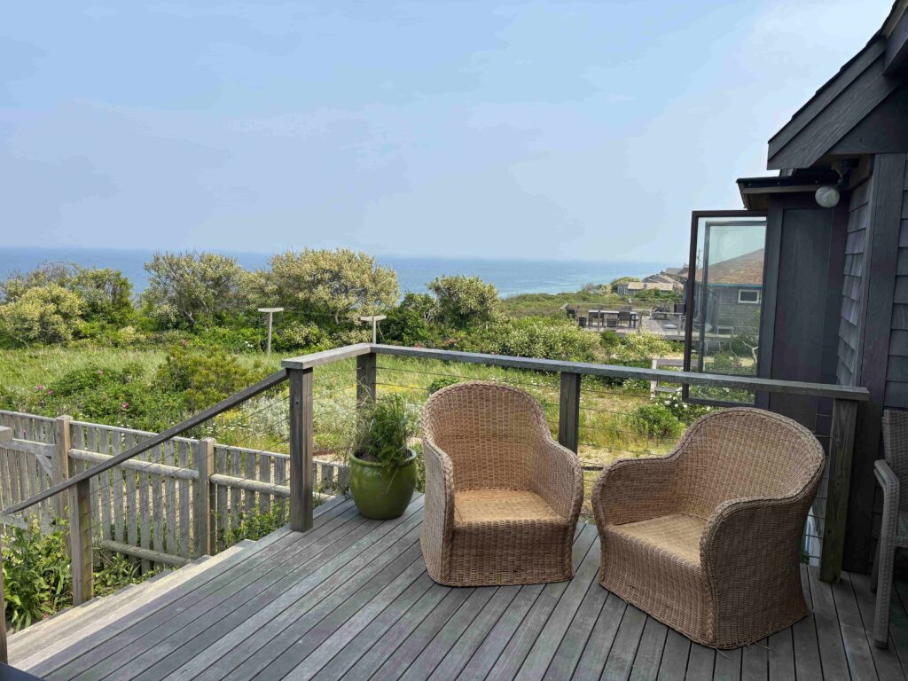 A spacious wooden deck with a flush threshold (visible in other photos) from the interior. The deck features a sturdy wooden railing and a wide, level surface for seating. Two wicker armchairs are positioned with plenty of surrounding space for a wheelchair or walker to turn. The background shows a view of the ocean.