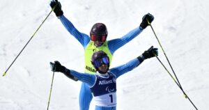 Two Paralympic alpine skiers in blue racing suits raise their arms in celebration on a snowy slope at the Milano Cortina 2026 Winter Games.