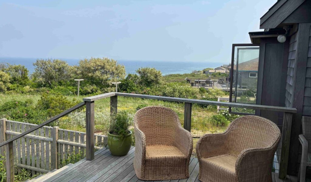 A spacious wooden deck with a flush threshold (visible in other photos) from the interior. The deck features a sturdy wooden railing and a wide, level surface for seating. Two wicker armchairs are positioned with plenty of surrounding space for a wheelchair or walker to turn. The background shows an unobstructed view of the ocean.