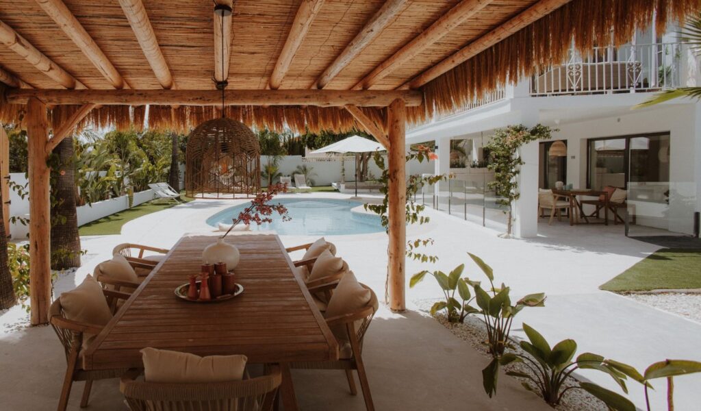 A luxurious outdoor dining area under a rustic, thatched wooden pergola at an accessible Mediterranean vacation villa, featuring a long wooden table on a smooth, level-access white tile floor that leads seamlessly to a turquoise swimming pool and a modern white-washed house surrounded by palm trees.