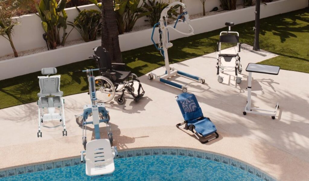 A collection of adaptive mobility and accessibility equipment arranged on a pool deck beside a tiled swimming pool. The equipment includes a manual wheelchair, a patient lift/hoist with a sling, a pool access chair (designed to lower users into the water), a shower/commode chair on wheels, a reclining pool access lounger with blue fabric, and a standing support frame. Green grass and palm trees are visible in the background, suggesting a warm-weather recreational or therapeutic facility.Sonnet 4.6