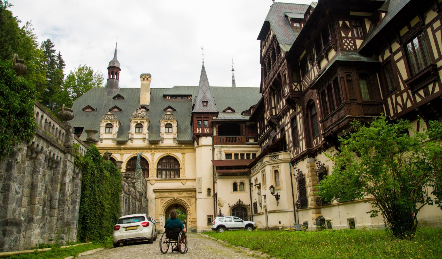 A photograph shows a person in a manual wheelchair from behind, centered on a cobblestone path leading toward an ornate, multi-story castle or palace. The building features complex architecture with a mix of cream-colored stone walls, dark timber framing, and steeply pitched slate-gray roofs with decorative turrets and dormer windows. To the left, a tall stone wall covered in green ivy runs alongside the path. Two white cars are parked nearby—one just ahead of the person in the wheelchair and another further back near the building's entrance archway. The scene is set under an overcast sky, surrounded by lush green trees and a small grassy area to the right.