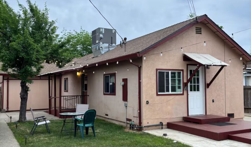 A small backyard with a detached stucco building featuring a light tan exterior and red trim. A white door with a small awning sits above a low wooden platform deck with a ramp. There is a small outdoor table with chairs on a patch of grass.