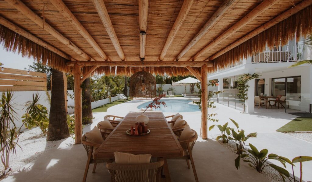 A luxurious outdoor dining area under a rustic, thatched wooden pergola at an accessible Mediterranean vacation villa, featuring a long wooden table on a smooth, level-access white tile floor that leads seamlessly to a turquoise swimming pool and a modern white-washed house surrounded by palm trees.