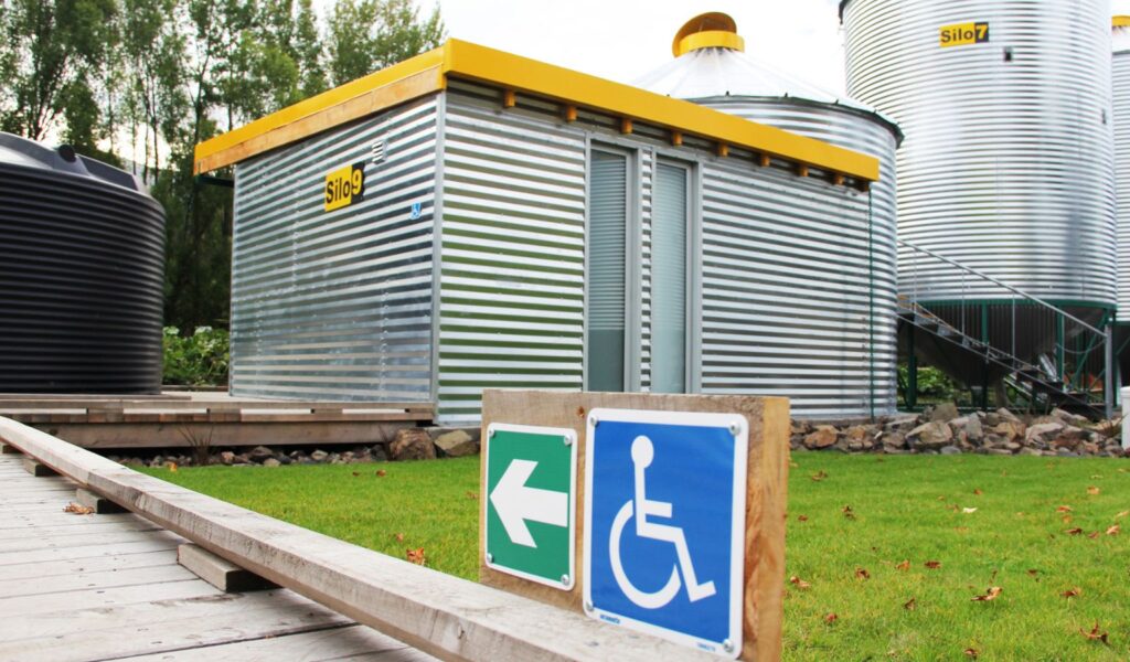 Exterior view of Silo 9 at SiloStay, showing a corrugated metal accessible unit with a yellow trim, level wooden boardwalk, and a clear blue and white wheelchair accessibility sign in the foreground.
