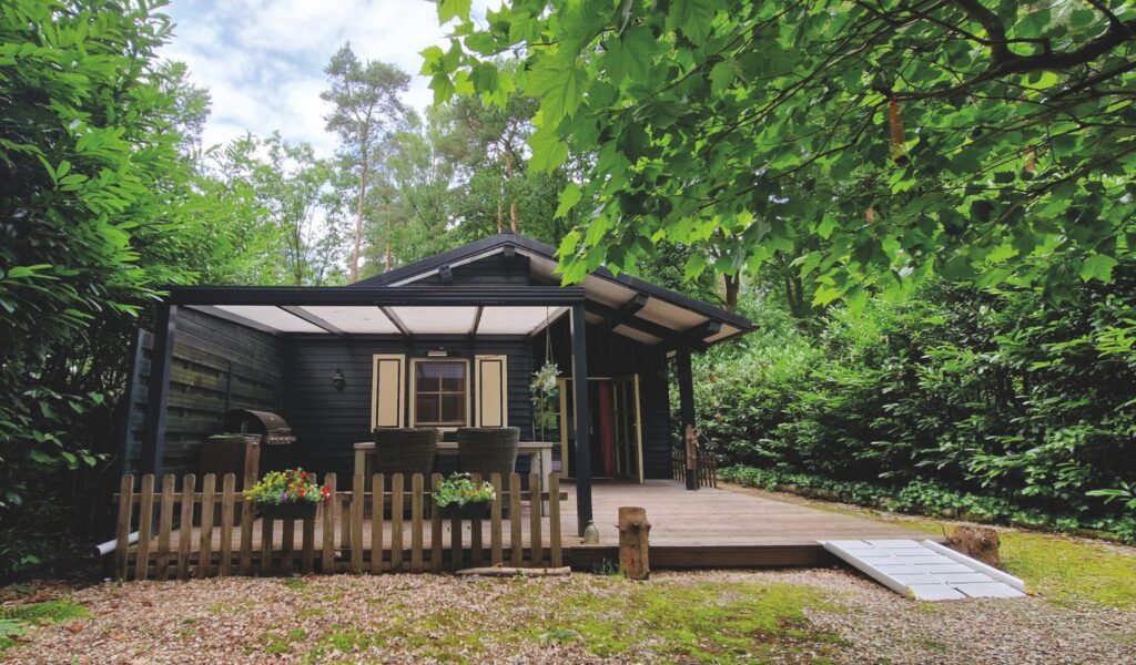 Accessible rental property in Limburg, Belgium — a charming dark-painted wooden chalet nestled among lush green trees, featuring a covered outdoor deck with patio furniture, a BBQ grill, colorful flower boxes along a wooden picket fence, and a accessibility ramp leading to the entrance.
