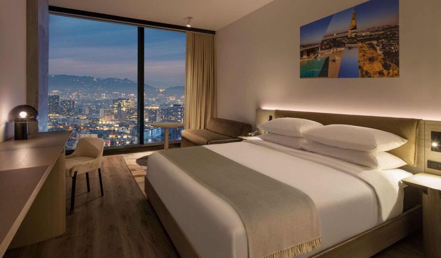 Spacious, modern hotel room at AC Hotel Downtown Los Angeles by Marriott with a king bed, hardwood floors and floor-to-ceiling windows overlooking the illuminated LA cityscape at night – accessible Los Angeles accommodation
