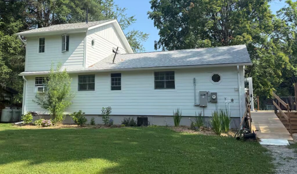 The exterior of a white, two-story siding house. A long, gradual wooden ramp with sturdy handrails leads from a gravel path up to the main entrance, providing step-free access to the home.