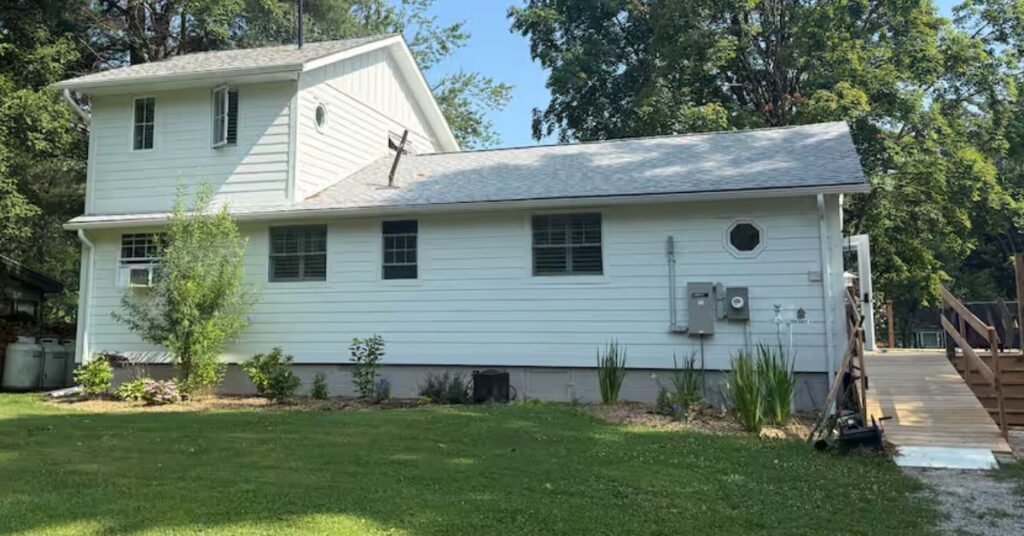 The exterior of a white, two-story siding house. A long, gradual wooden ramp with sturdy handrails leads from a gravel path up to the main entrance, providing step-free access to the home.