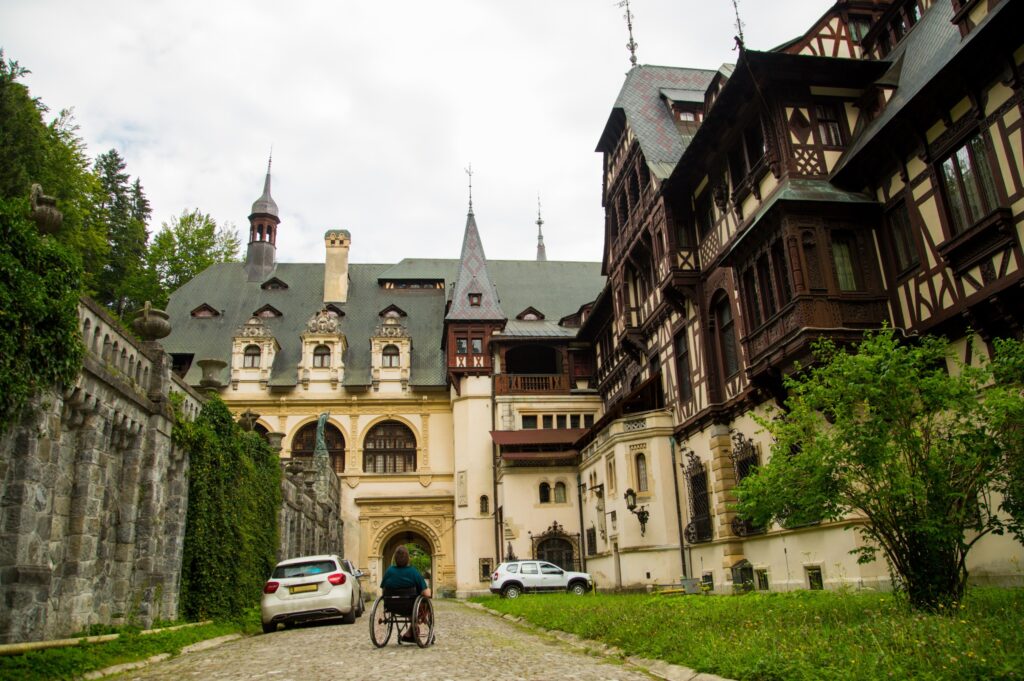 Wheelchair user approaching the entrance of Peleș Castle in Sinaia, Romania, along a cobblestone path through the private estate