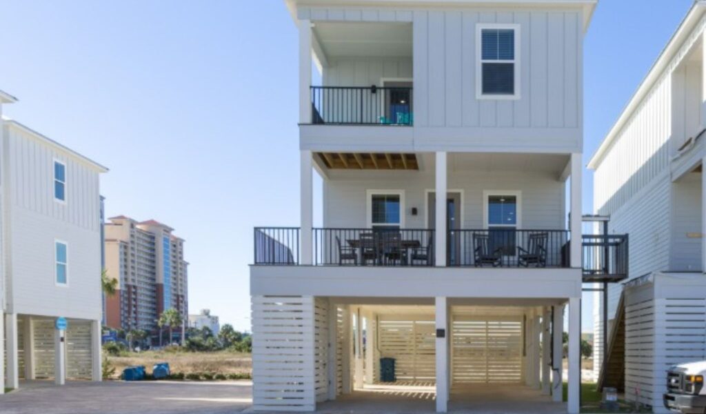 Exterior view of a white, three-story accessible vacation rental on stilts. Features include a paved, shaded carport and a visible external platform lift (elevator) connecting the ground to the upper balconies. A large, furnished second-floor deck is shown with an outdoor dining table and rocking chairs. Tall, multistory buildings are visible in the background.