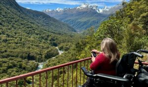 Wheelchair user in a power chair photographs the Hollyford Valley from an accessible viewpoint on the Milford Road, South Island New Zealand, with snow-capped peaks and a glacial river below