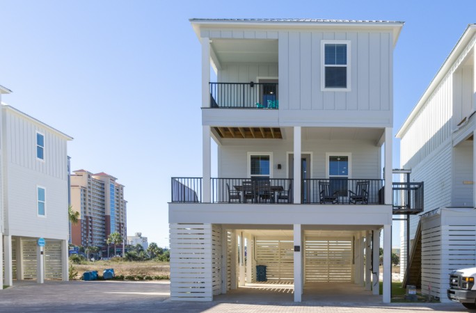 Exterior view of a white, three-story accessible vacation rental on stilts. Features include a paved, shaded carport and a visible external platform lift (elevator) connecting the ground to the upper balconies. A large, furnished second-floor deck is shown with an outdoor dining table and rocking chairs. Tall, multistory buildings are visible in the background.