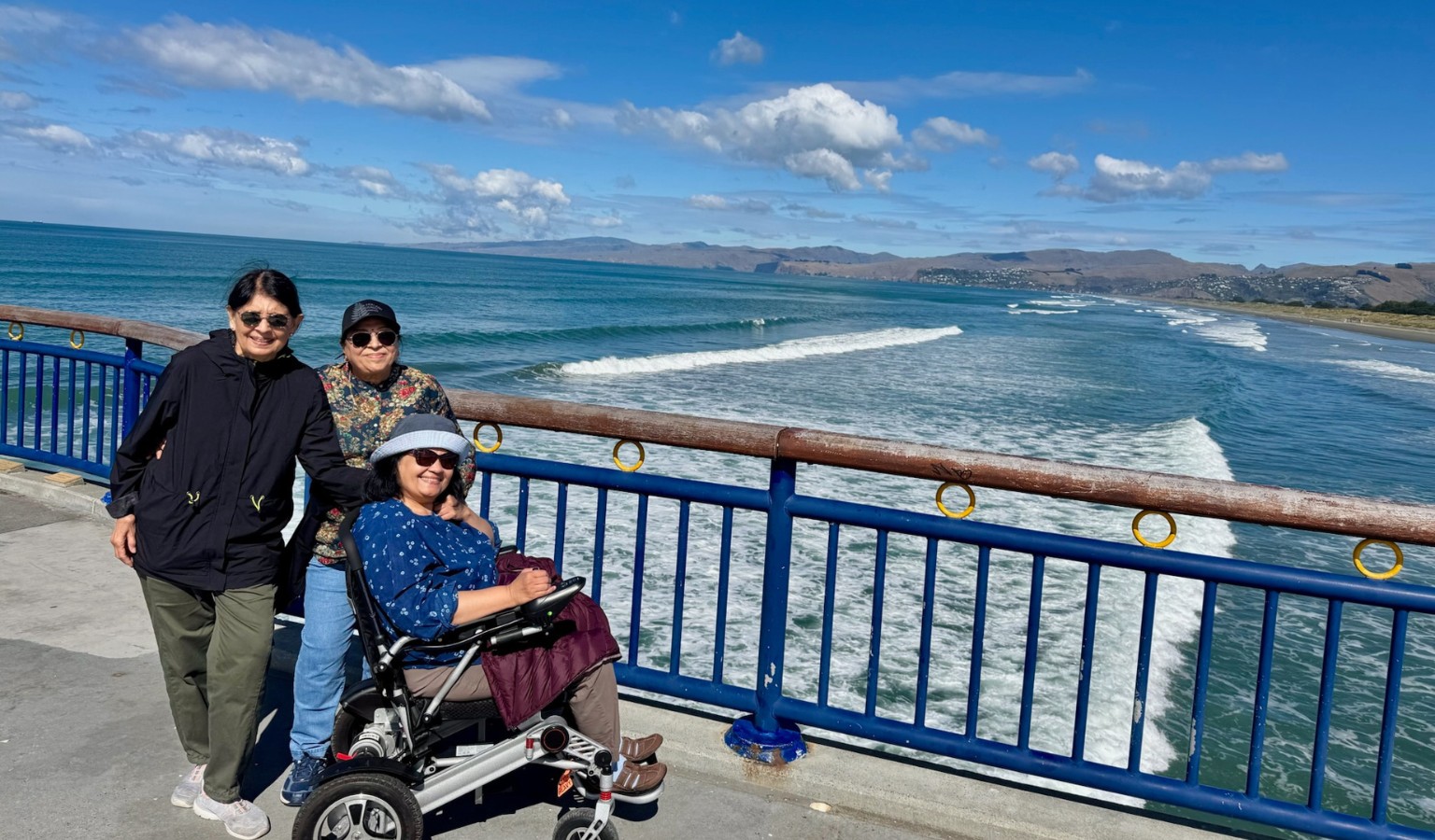 Three women on an accessible coastal pier in New Zealand, one using a folding travel wheelchair, with breaking waves and South Island hills behind them