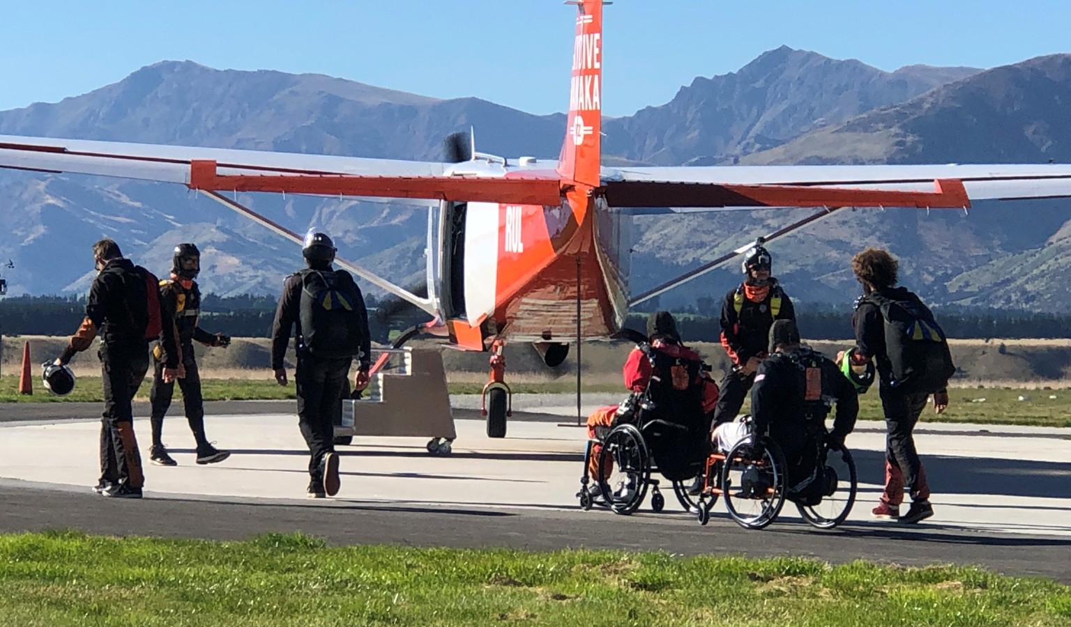 Wheelchair user in skydiving gear being assisted toward a Skydive Wanaka aircraft on the runway, with the Southern Alps visible behind, Queenstown region New Zealand