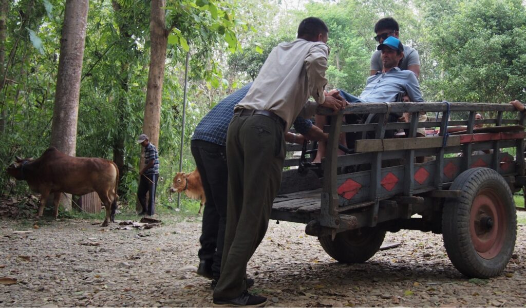 Jezza Williams, a disabled adventurer and tetraplegic, is helped onto a wooden wagon cart by local men on a rural path in Nepal, with cattle grazing in the background.