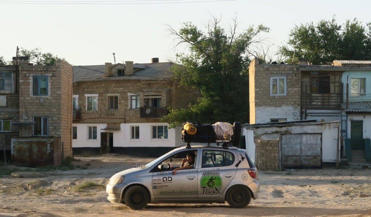 A small silver car branded with the Making Trax Foundation logo drives through a dusty Central Asian town, loaded with gear on the roof rack.