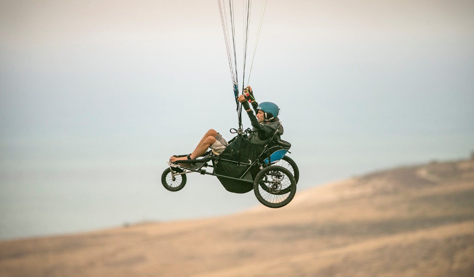Jezza Williams paraglides using a custom reclined three-wheeled rig suspended beneath a paraglider canopy, hands on the controls, over an open desert landscape.