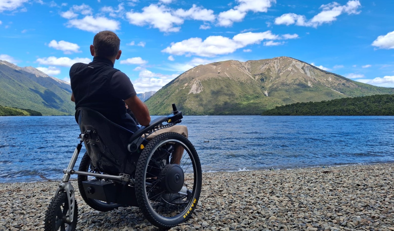 Jezza Williams sits in his all-terrain wheelchair on the pebbly shore of a lake in New Zealand's South Island, looking out toward mountain peaks under a bright blue sky.