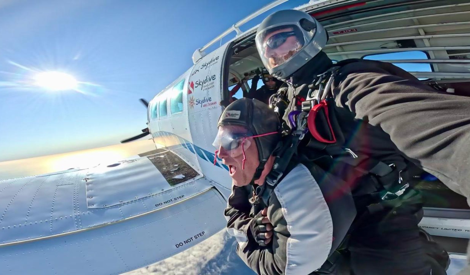 Jezza Williams tandem skydives over a snowy New Zealand landscape, mouth open in a shout, with a Skydive Franz Josef and Abel Tasman branded plane visible above.