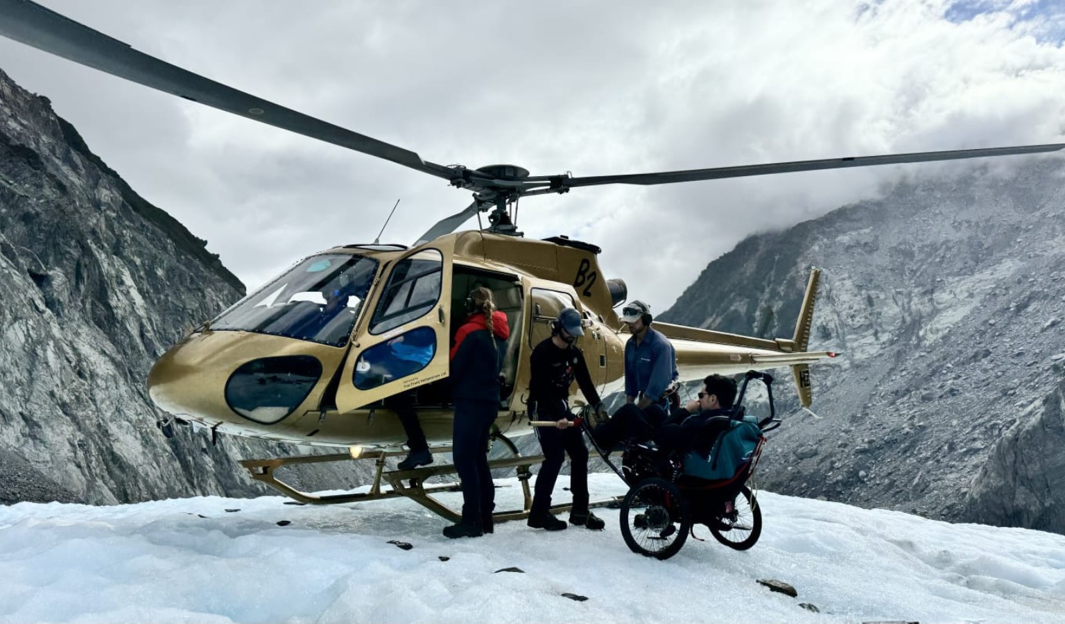 A wheelchair user in a specialised outdoor chair is assisted by guides beside a helicopter on a glacier, with dramatic snow-covered mountain walls rising behind them.