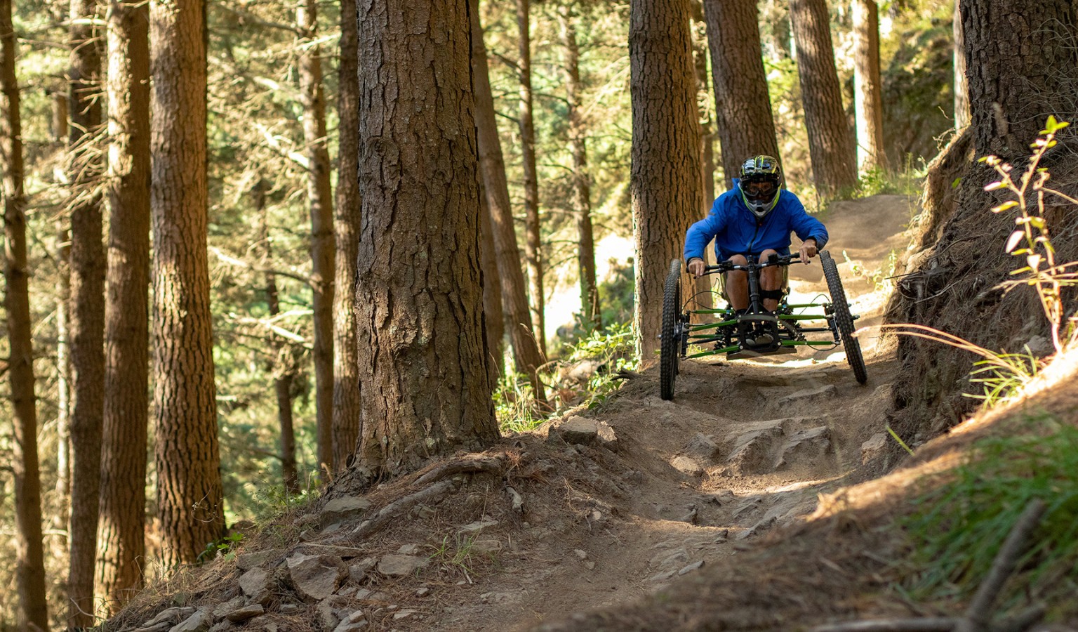 A rider on an adaptive quad bike descends a steep, rooted forest mountain bike trail at speed, wearing a blue jacket and full-face helmet.