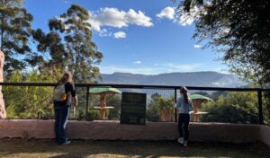 A mother and her young daughter stand at a zoo enclosure railing, looking out over a lush green landscape with mountains in the background, on a sunny day in Brazil.