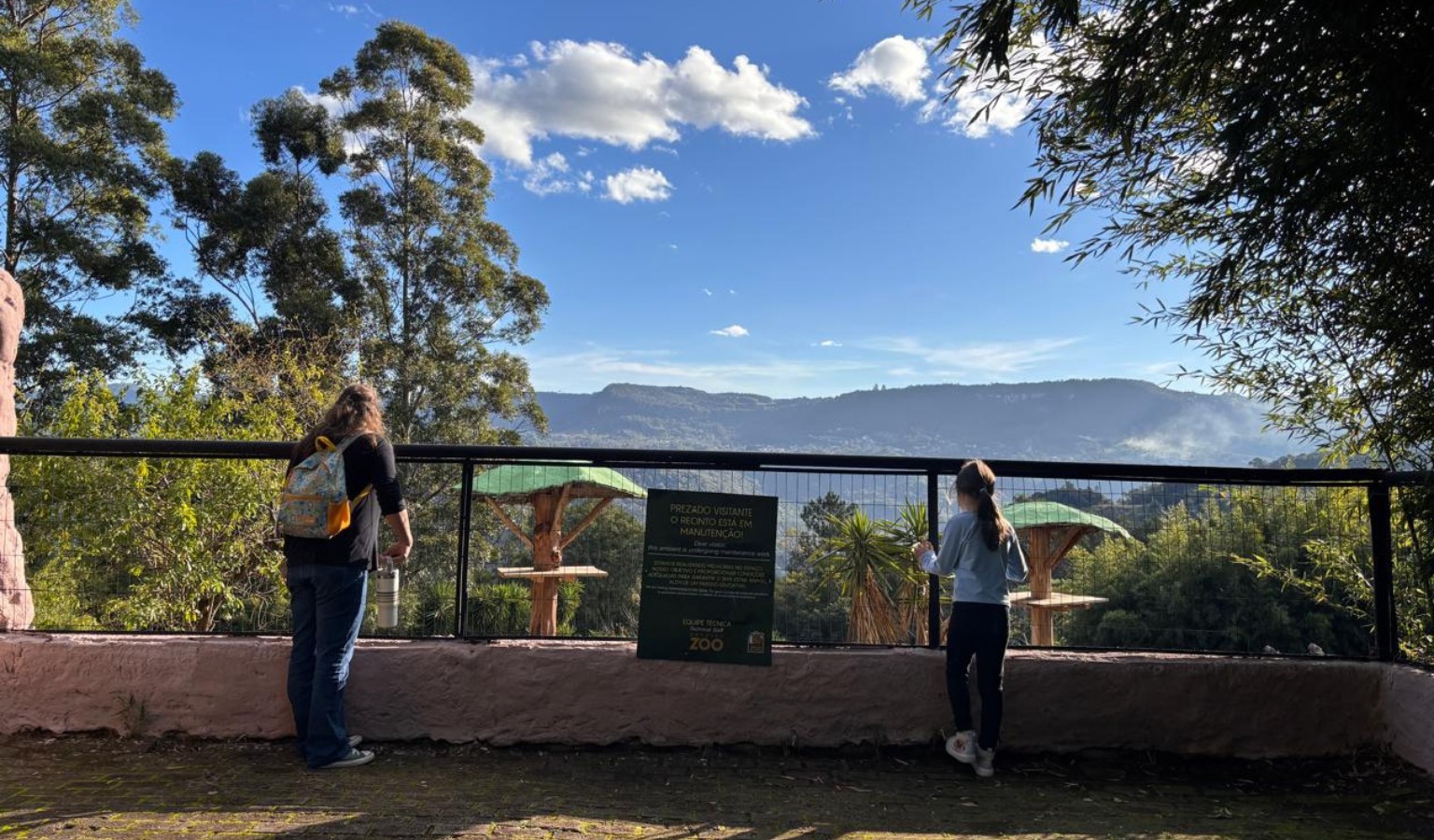 A mother and her young daughter stand at a zoo enclosure railing, looking out over a lush green landscape with mountains in the background, on a sunny day in Brazil.
