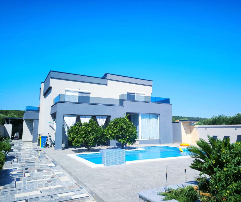 Modern accessible two-story white and gray villa in Bibinje, with large windows and glass balconies overlooking a rectangular outdoor swimming pool. A tiled patio surrounds the pool, with lounge chairs along a privacy wall, leafy trees and tropical plants in the foreground, and a clear blue sky above.