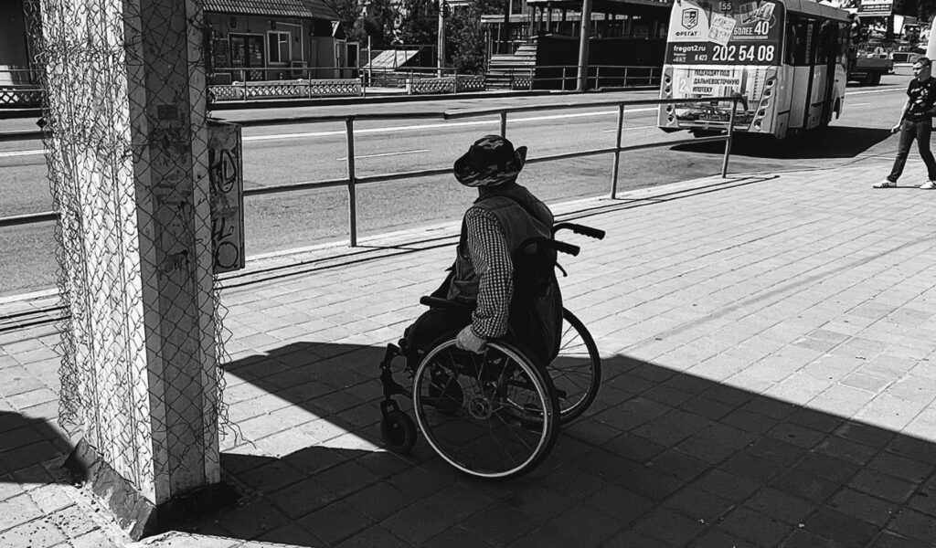 Black and white photo of a wheelchair user waiting alone at the edge of a sidewalk, separated from a passing city bus by a long metal railing — a quiet picture of navigating a world not built with him in mind.