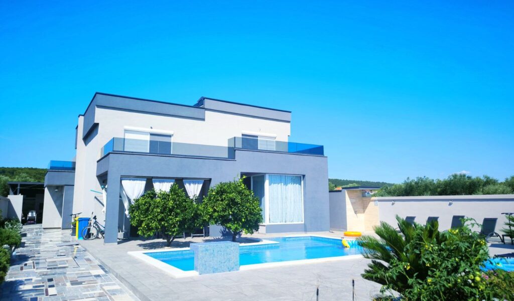 Modern accessible two-story white and gray villa in Bibinje, with large windows and glass balconies overlooking a rectangular outdoor swimming pool. A tiled patio surrounds the pool, with lounge chairs along a privacy wall, leafy trees and tropical plants in the foreground, and a clear blue sky above.