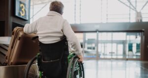 A wheelchair user waits beside a suitcase in an airport baggage reclaim area, facing toward the terminal exit.