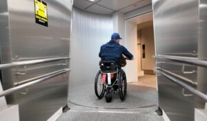 Shane Hryhorec wheels through an airport jet bridge in his manual wheelchair, seen from behind, with a black SmartDrive power-assist device attached beneath the seat and a red "gate" tag hanging from the frame.