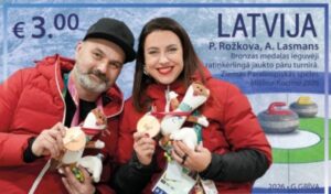 Latvian €3 postage stamp showing curlers P. Rožkova and A. Lasmans in red team jackets, smiling and holding up their bronze medals and Games mascot toys. Latvian text notes their bronze in wheelchair curling mixed doubles at the Milan–Cortina 2026 Winter Paralympics; curling stones sit on ice at right.