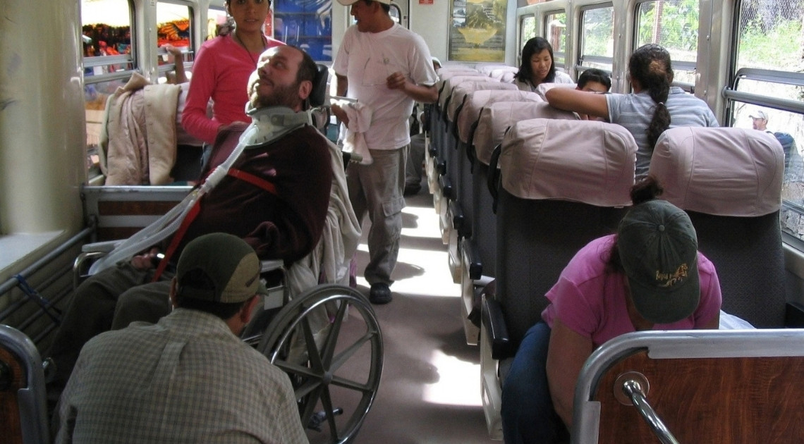 Inside a train carriage, a man sits in a manual wheelchair secured with red straps, wearing a neck brace with a breathing tube attached. An assistant kneels beside the chair while other passengers sit in rows of seats and a staff member stands in the aisle.