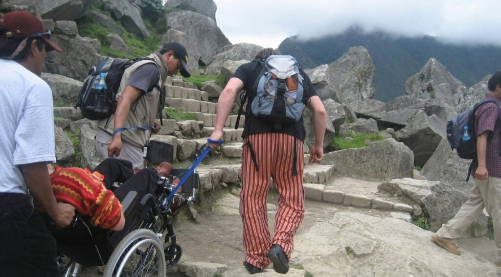 A wheelchair user wearing a bright red and yellow knit sweater is pulled backward up a flight of stone steps at Machu Picchu by a team of assistants. One man walks ahead holding a blue strap attached to the chair, while others support it from the sides and behind. Large granite boulders surround the path, with cloud-covered mountains in the background.