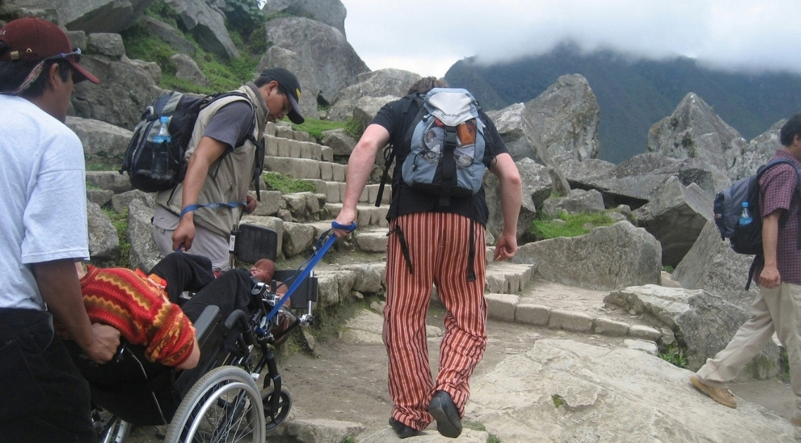 A wheelchair user wearing a bright red and yellow knit sweater is pulled backward up a flight of stone steps at Machu Picchu by a team of assistants. One man walks ahead holding a blue strap attached to the chair, while others support it from the sides and behind. Large granite boulders surround the path, with cloud-covered mountains in the background.