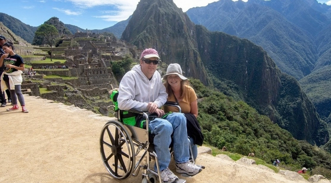 A smiling man in a manual wheelchair, fitted with green padded supports, sits beside a smiling woman crouched next to him at a viewpoint above Machu Picchu. The terraced stone ruins of the citadel and the steep green peak of Huayna Picchu rise behind them under a blue sky.
