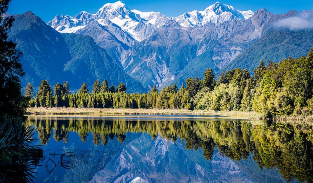 Snow-capped Aoraki/Mount Cook and Mount Tasman reflected in the still water of Lake Matheson on New Zealand's West Coast, with native forest lining the far shore.