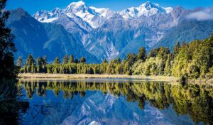 Snow-capped Aoraki/Mount Cook and Mount Tasman reflected in the still water of Lake Matheson on New Zealand's West Coast, with native forest lining the far shore.