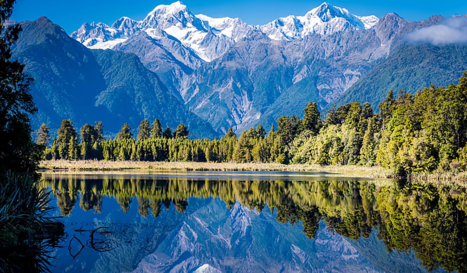 Snow-capped Aoraki/Mount Cook and Mount Tasman reflected in the still water of Lake Matheson on New Zealand's West Coast, with native forest lining the far shore.