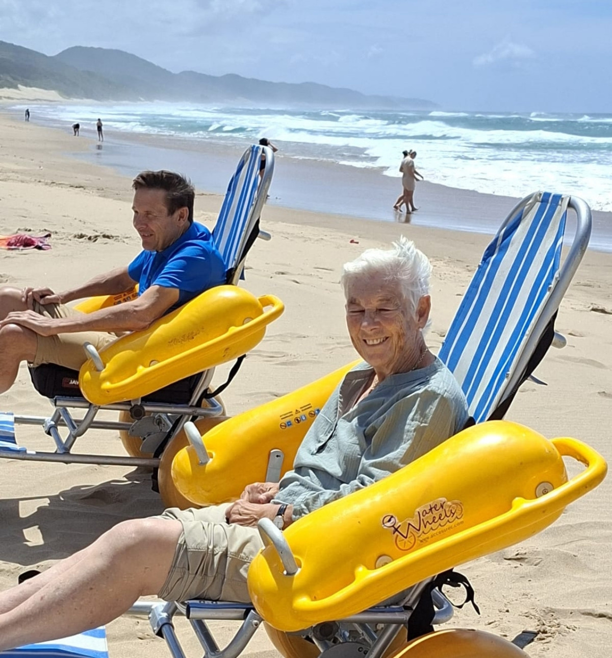Two people sit in yellow floating beach wheelchairs on a sandy beach, a smiling white-haired woman in front, with waves and green hills behind them.