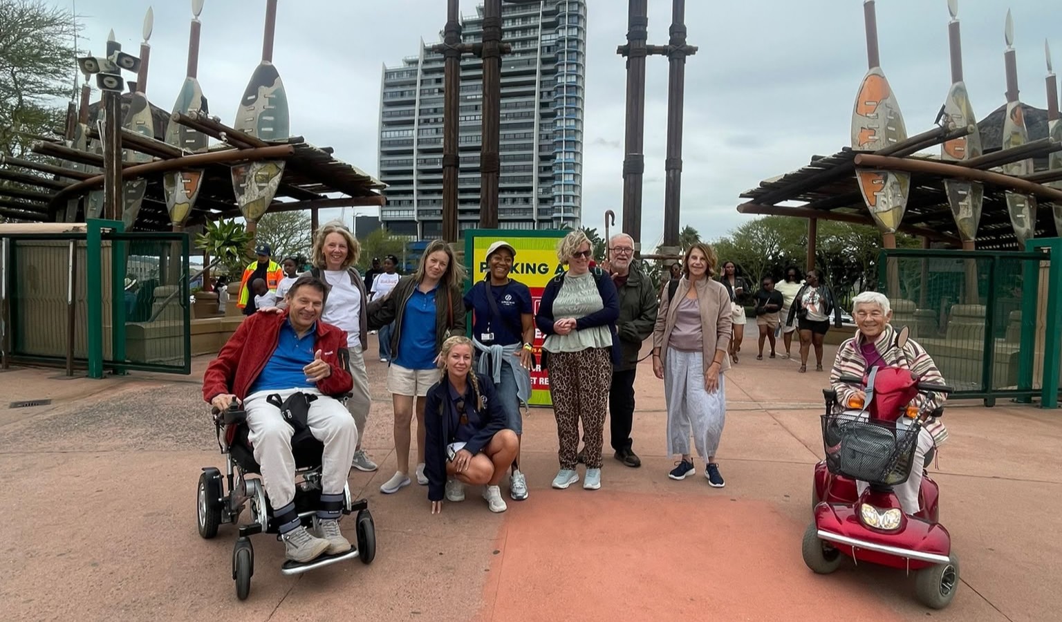 A group of nine people smile outside the entrance to uShaka Marine World in Durban, including a man in a power wheelchair giving a thumbs up and a woman on a red mobility scooter. Tall Zulu-style shield and spear sculptures frame the entrance behind them.
